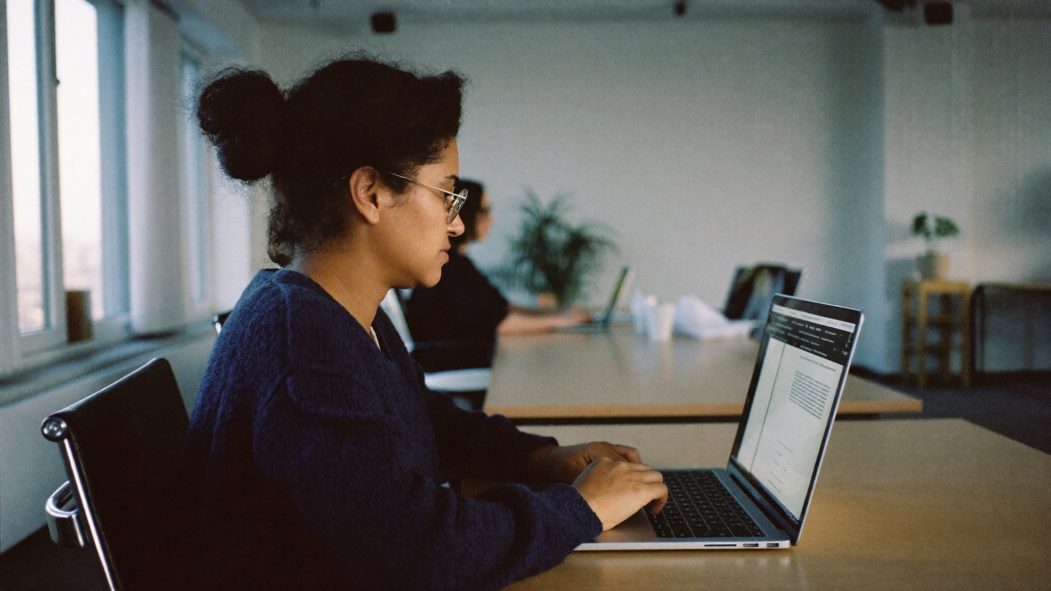 Adriana, coordenadora de RH, digitando no notebook numa mesa comprida de escritório, com uma colega trabalhando ao fundo